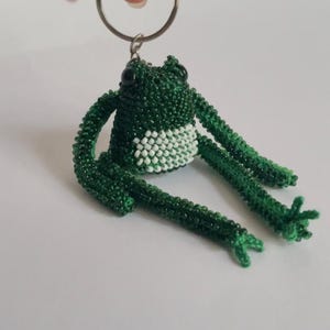 May include: A green beaded frog keychain with white beads on its belly. The frog is sitting with its legs outstretched and has black bead eyes.