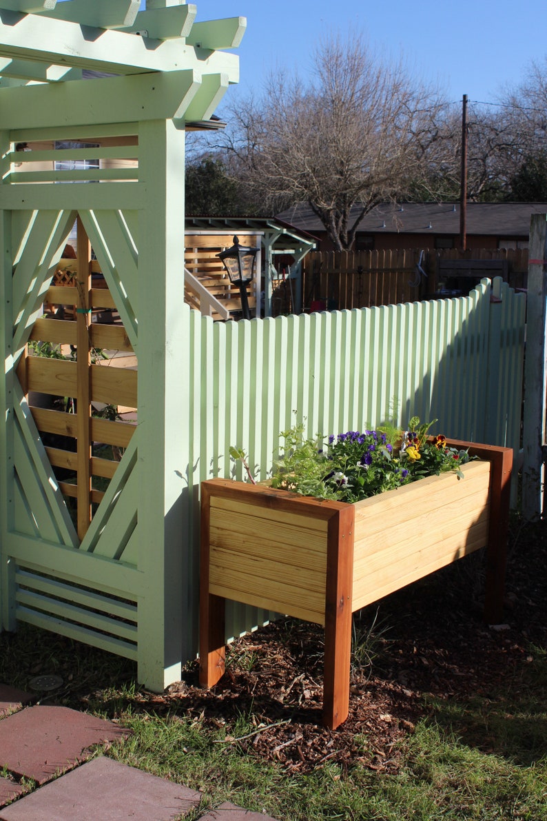 a modern planter sits outside against an arbor, filled with green plants and flowers it shows it can eliminate the need to bend over and ache your back when weeding and planting