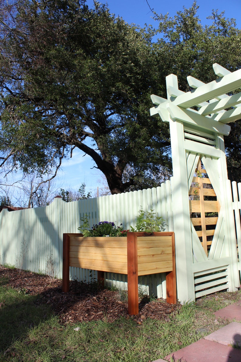 The modern planter, with horizontal pine slats, all the way around, are cornered by the redish brown hue of cedar. It shows that the plans for this garden bed can be a great accent to arbors