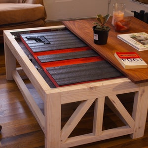 Puede incluir: Una mesa de centro de madera con un compartimento oculto que se abre para revelar un arma de fuego y un forro rojo y negro. La mesa tiene un acabado pintado de blanco y un diseño sencillo.