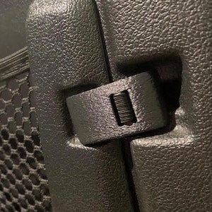 May include: Close-up of a black plastic latch with a black elastic strap. The latch is attached to a black mesh panel.