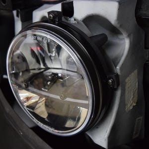 May include: A close-up of a round, black, and silver car headlight with a clear lens. The headlight has a black bezel and a silver trim ring. The headlight is mounted on a white car body. The text "DOT SAE" is visible on the headlight.