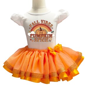 Pode incluir: Um body branco de manga curta com um gráfico de uma abóbora, um arco-íris e o texto FALL VIBES and PUMPKIN SPICE, combinado com uma saia tutu de tule laranja brilhante com um laço de cetim amarelo e uma borda dourada com glitter.