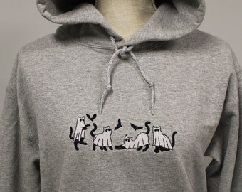 Puede incluir: Una sudadera con capucha de color gris jaspeado que presenta un diseño bordado de cuatro gatos vestidos con disfraces de fantasma blancos. Los gatos aparecen en varias posturas con pequeños murciélagos negros volando sobre ellos. Incluye un cordón gris.