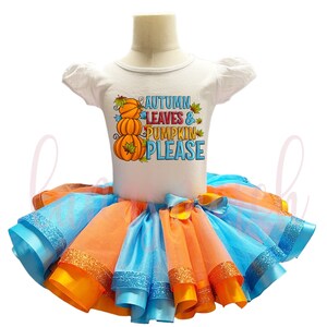 Pode incluir: Um body branco de manga curta com um gráfico de três abóboras laranja empilhadas e o texto AUTUMN LEAVES & PUMPKIN PLEASE, combinado com uma saia tutu de tule multicamadas em tons alternados de azul brilhante, laranja e amarelo.