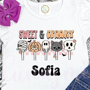 Könnte beinhalten: Ein weißes Kurzarm-T-Shirt mit einem Aufdruck, der den Text "SWEET & SPOOKY" über fünf Halloween-Cake-Pops in Form von Mumie, Kürbis, Geist, schwarzer Katze und Totenkopf zeigt, mit dem Namen "Sofia" in schwarzer Schrift darunter.