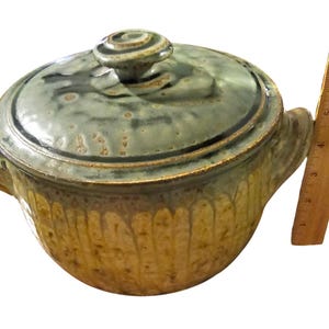 May include: A ceramic lidded casserole dish with a green and tan glaze. The dish has two handles and a spiral knob on the lid. A wooden ruler is next to the dish, measuring approximately 6 inches.