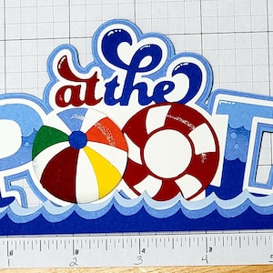 Pode incluir: Corte de papel azul e branco das palavras "at the POOL" com um salva-vidas vermelho e branco e uma bola de praia multicolorida.