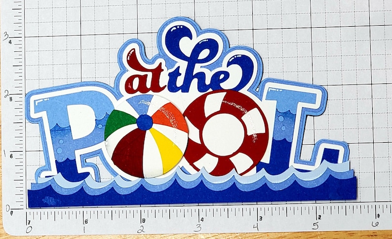 Pode incluir: Corte de papel azul e branco das palavras "at the POOL" com um salva-vidas vermelho e branco e uma bola de praia multicolorida.