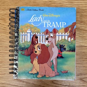 May include: A spiral-bound Little Golden Book titled Walt Disney's Lady and the Tramp. The cover features a colorful illustration of Lady, Tramp, Trusty, and Jock in a grassy yard with a white picket fence in the background under a blue sky.