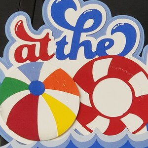 Pode incluir: Uma arte de papel azul e branco com as palavras "at the" em letras vermelhas e azuis. A arte apresenta um salva-vidas vermelho e branco e uma bola de praia vermelha, branca, amarela e verde. A arte &eacute; colocada sobre um padr&atilde;o de ondas azul e branco.