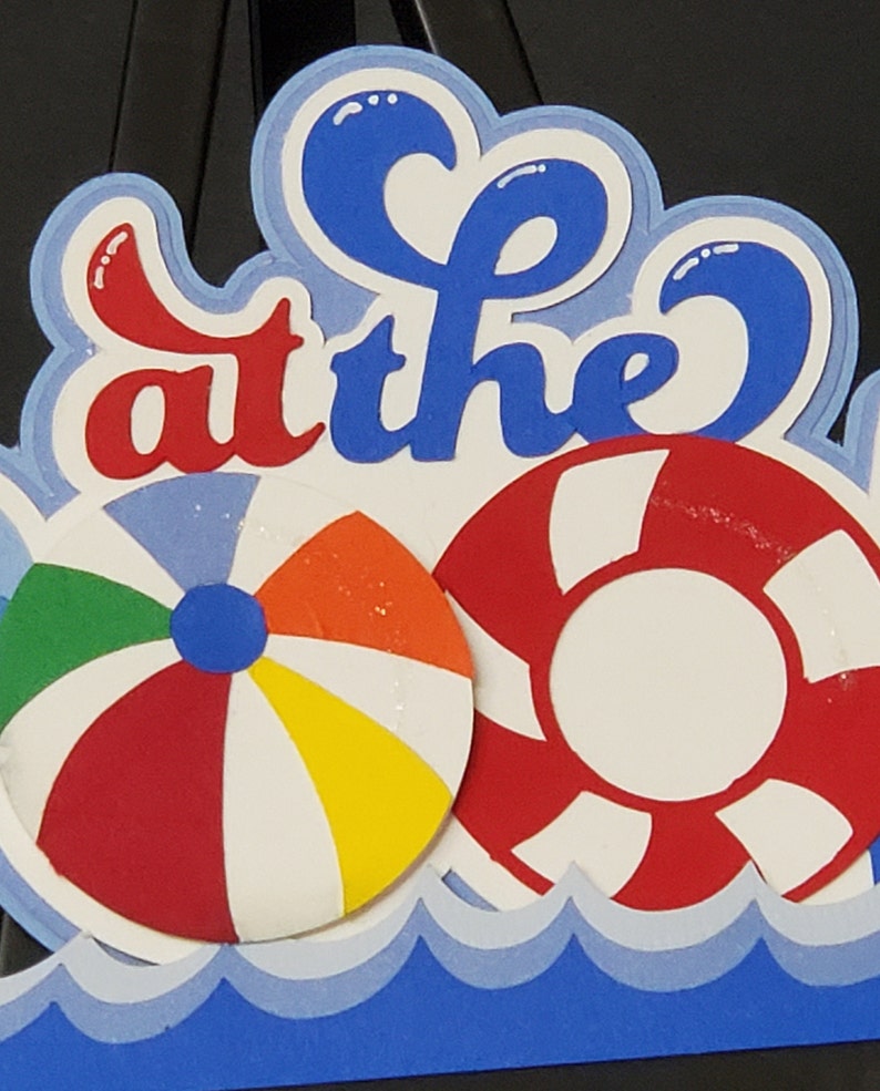 Pode incluir: Uma arte de papel azul e branco com as palavras "at the" em letras vermelhas e azuis. A arte apresenta um salva-vidas vermelho e branco e uma bola de praia vermelha, branca, amarela e verde. A arte &eacute; colocada sobre um padr&atilde;o de ondas azul e branco.