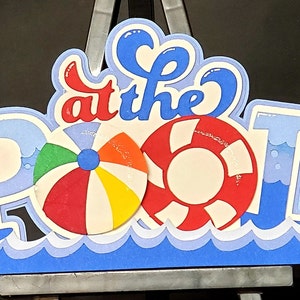 Pode incluir: Um sinal de papel azul e branco que diz "at the Pool" com uma bola de praia vermelha, branca e azul e um salva-vidas vermelho e branco.