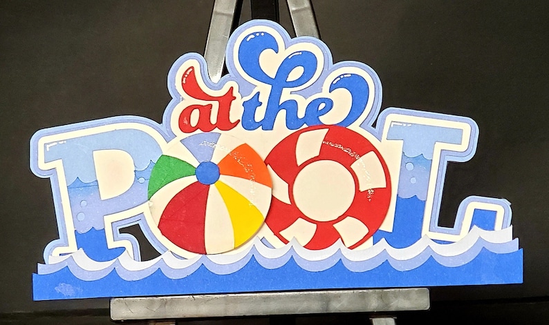 Pode incluir: Um sinal de papel azul e branco que diz "at the Pool" com uma bola de praia vermelha, branca e azul e um salva-vidas vermelho e branco.
