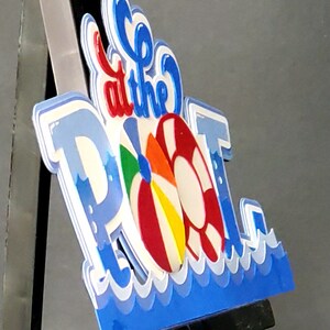 Pode incluir: Uma placa 3D azul e branca que diz "Go to the Pool" com uma bola de praia colorida e um salva-vidas vermelho e branco.