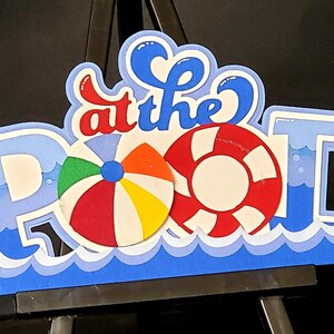 Pode incluir: Recorte de papel azul e branco das palavras "at the pool" com um salva-vidas vermelho e branco, uma bola de praia colorida e ondas azuis na parte inferior.