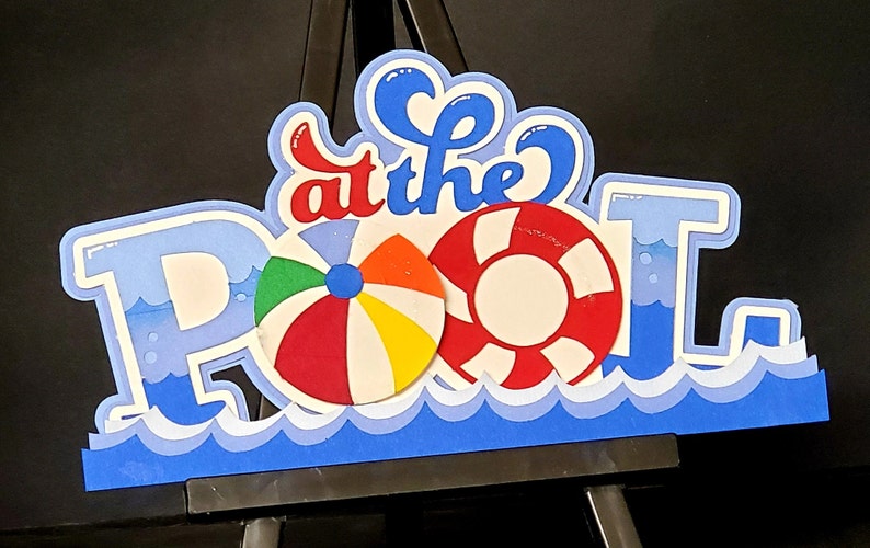 Pode incluir: Recorte de papel azul e branco das palavras "at the pool" com um salva-vidas vermelho e branco, uma bola de praia colorida e ondas azuis na parte inferior.