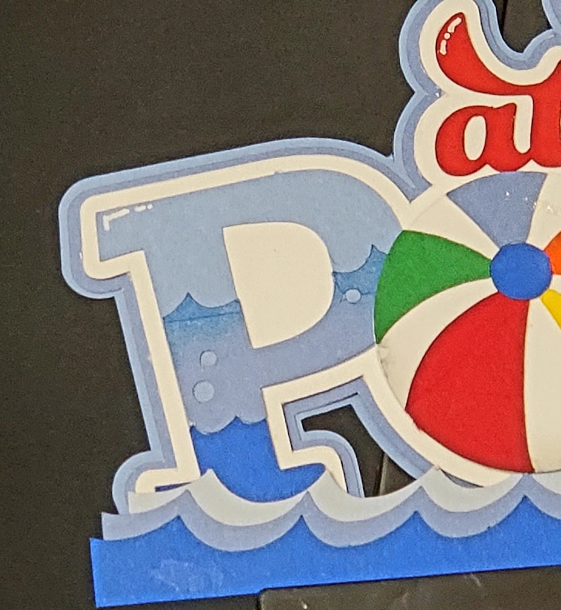 Pode incluir: Uma letra "P" azul e branca com uma bola de praia e ondas. A letra faz parte de uma palavra maior, possivelmente "Piscina" ou "Festa".