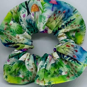 May include: A fabric scrunchie with a whimsical forest print. The design features green foliage, mushrooms, and berries in shades of green, blue, and purple. The scrunchie is a hair accessory.