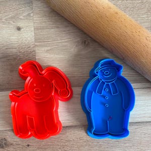 May include: A red plastic cookie cutter shaped like a dog with long ears and a blue plastic cookie cutter shaped like a snowman wearing a hat and scarf, positioned next to a wooden rolling pin. The cutters feature raised internal details.