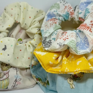 Puede incluir: Tres scrunchies de tela apilados. El scrunchie superior es blanco con un patrón floral en tonos de azul, verde y rosa. El scrunchie del medio es amarillo con un patrón floral blanco. El scrunchie inferior es azul claro con un patrón floral blanco y una pequeña ilustración de conejo.