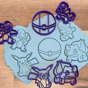May include: Purple plastic cookie cutters featuring cartoon characters and a Poké Ball. The cutters are arranged on a light blue surface, with the impressions of the characters visible.
