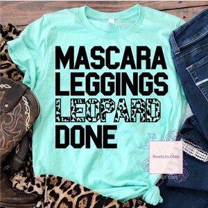 May include: A turquoise blue t-shirt with black text that reads "Mascara Leggings Leopard Done". The word "Leopard" is printed in a leopard print pattern.