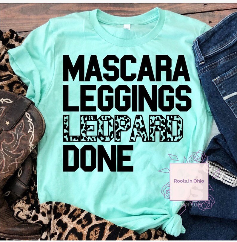 May include: A turquoise blue t-shirt with black text that reads "Mascara Leggings Leopard Done". The word "Leopard" is printed in a leopard print pattern.