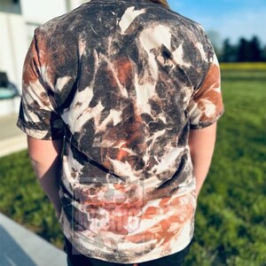 Może przedstawiać: Koszulka tie-dye w kolorze brązowym, czarnym i białym z kr&oacute;tkim rękawem i okrągłym dekoltem.