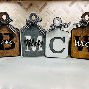 以下が含まれることがあります： 黒と白のチェック柄のリボンが付いた木製キッチンサインが4つ。それぞれのサインには、大きなイニシャルと下に名前が書かれています。Baker、Metz、C、Wickです。