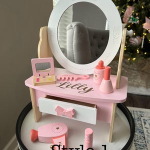 May include: Pink wooden vanity set with a mirror, a drawer, and a comb. The vanity has a pink bow on the top and the name "Lilly" is written on the front. The set includes a pink hair dryer and a pink lipstick.