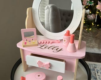 May include: Pink wooden vanity set with a mirror, a drawer, and a comb. The vanity has a pink bow on the top and the name "Lilly" is written on the front. The set includes a pink hair dryer and a pink lipstick.