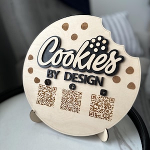 May include: A wooden sign in the shape of a cookie with a bite taken out. The sign reads "Cookies BY DESIGN" in black lettering. Three QR codes are printed below the text. The cookie has brown chocolate chip details.