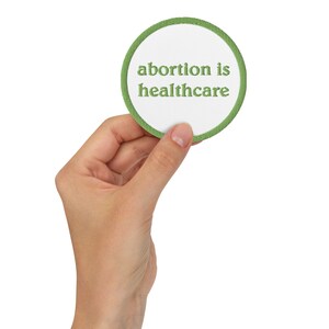 Könnte beinhalten: Ein runder grüner und weißer Aufnäher mit dem Text "abortion is healthcare", der in Grün gestickt ist.