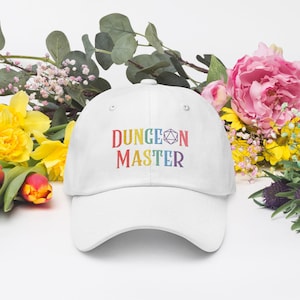 May include: A white baseball cap with the text "DUNGEON MASTER" embroidered in rainbow colors. The cap features a curved brim and a six-sided die symbol. The background includes colorful flowers and greenery.
