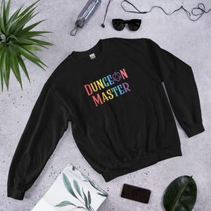 May include: Black sweatshirt with the text "DUNGEON MASTER" in rainbow colors. The design includes a multi-sided die graphic. The sweatshirt is a crew neck style and is displayed on a neutral surface.