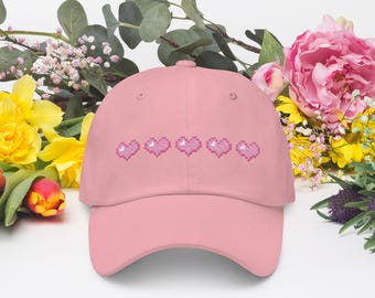 Può includere: Un cappellino da baseball rosa chiaro con una fila di cinque cuori pixelati ricamati in una tonalità di rosa più scura sul pannello frontale. Il cappellino presenta una visiera curva e una corona strutturata dal design semplice.