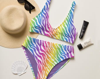 May include: A two-piece swimsuit with a vibrant rainbow zebra print. The top is a triangle style, and the bottoms are high-waisted. Accessories include a straw hat, sunglasses, sunscreen, and a seashell.