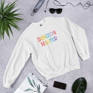 May include: White sweatshirt with the text "DUNGEON MASTER" in rainbow colors. The design includes a D20 die graphic. A casual, long-sleeved garment for fans of tabletop role-playing games.