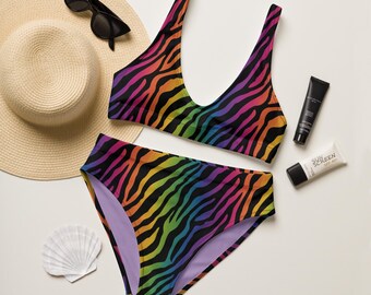 May include: A two-piece swimsuit with a vibrant rainbow zebra print. The top has a scoop neck, and the bottoms are high-waisted. Accessories include sunglasses, a straw hat, sunscreen, and a seashell.