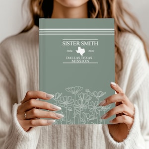 Op de afbeelding: Een saliegroen notitieboek met de tekst "SISTER SMITH" en "DALLAS TEXAS MISSION". De omslag heeft een witte Texas-omtrek en bloemenillustraties. De jaren 2024 en 2026 worden ook weergegeven.
