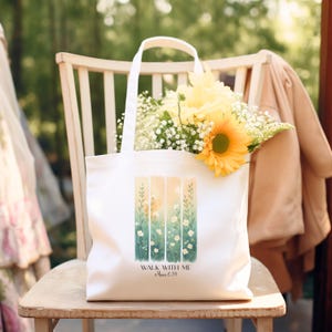 May include: A white canvas tote bag featuring a vertical watercolor-style print of green stems and small white flowers. Below the illustration, the text reads "WALK WITH ME" followed by "Micah 6:8" in a dark, elegant serif font.