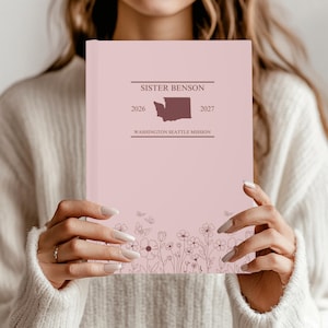 Op de afbeelding: Een roze notitieboek met de tekst "SISTER BENSON" en "WASHINGTON SEATTLE MISSION". Het notitieboek heeft een bloemenontwerp aan de onderkant en een kaart van de staat Washington. De jaren 2026 en 2027 staan ook op de omslag.
