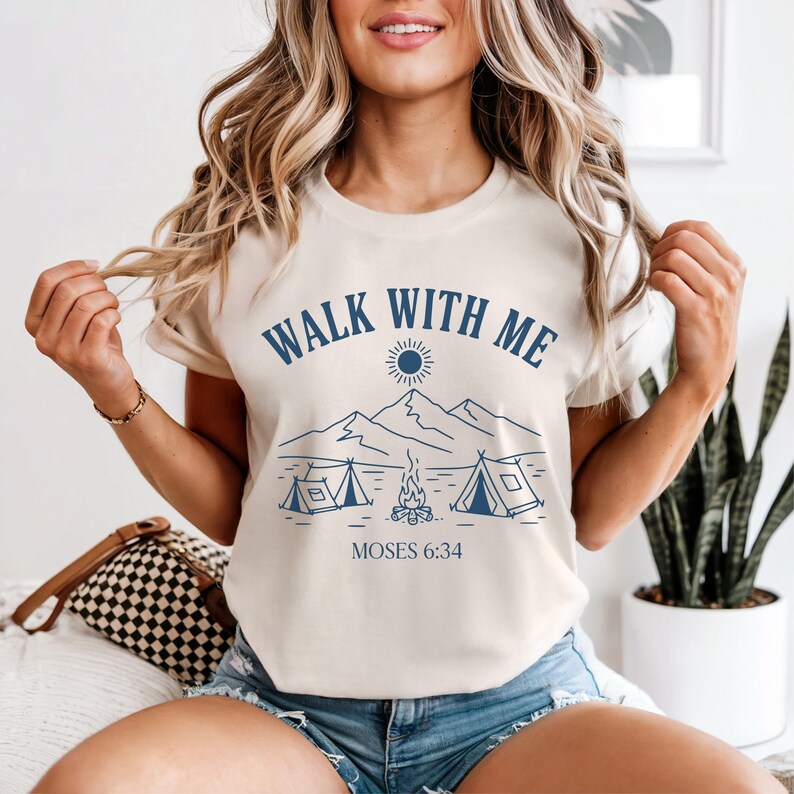 May include: Cream-colored t-shirt with the text "WALK WITH ME" in navy blue, along with a graphic of mountains, tents, a campfire, and the text "MOSES 6:34". The shirt has short sleeves.