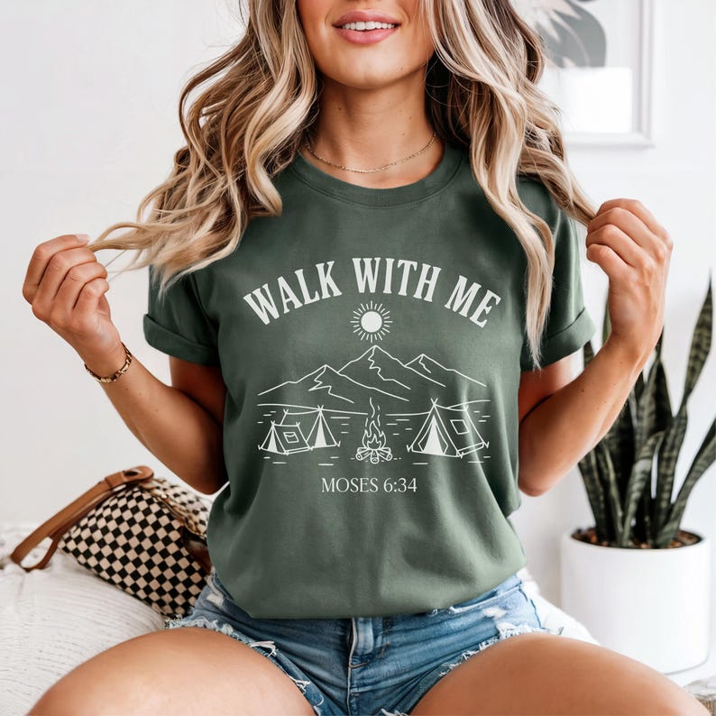 May include: Olive green t-shirt with the text "WALK WITH ME" above a graphic of mountains, tents, and a campfire. The text "MOSES 6:34" is below the graphic. The shirt has short sleeves and a crew neck.