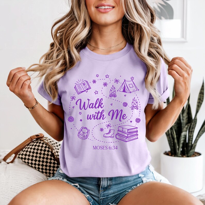 Pu&ograve; includere: T-shirt lilla chiaro con un design grafico viola. Il design include il testo "Walk with Me" e illustrazioni di un libro, una tenda, un fal&ograve;, uno zaino e stivali. &Egrave; incluso anche il testo "MOSES 6:34".