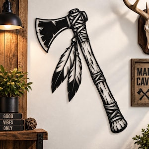 May include: A black and white metal wall art piece shaped like a tomahawk axe. The design features a textured handle wrapped in cordage, a sharp blade with a notched edge, and two dangling feathers attached to the head of the axe with a small bead.