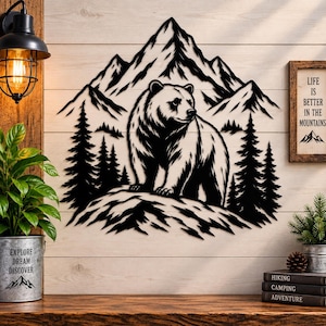 Puede incluir: Una pieza de arte mural de metal negro que muestra un oso de pie frente a una cordillera y pinos. A la derecha, un letrero enmarcado muestra el texto: LIFE IS BETTER IN THE MOUNTAINS con un pequeño gráfico de montaña en la parte inferior.