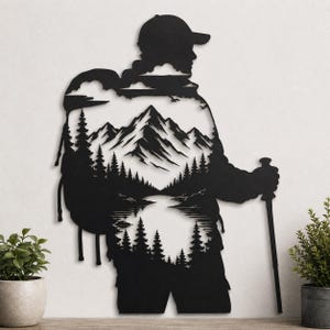 May include: A black metal wall art piece shaped as a silhouette of a hiker wearing a cap and carrying a large backpack. The interior of the figure features a detailed cut-out landscape of snow-capped mountains, pine trees, and a calm lake.