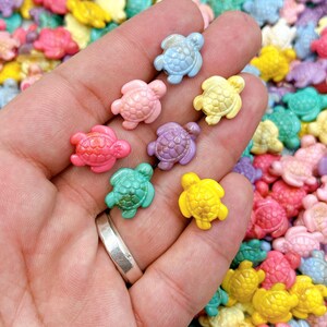 May include: A hand holding a collection of colorful turtle-shaped beads in various shades of pink, blue, green, yellow, and purple. The beads are small and appear to be made of a smooth material.